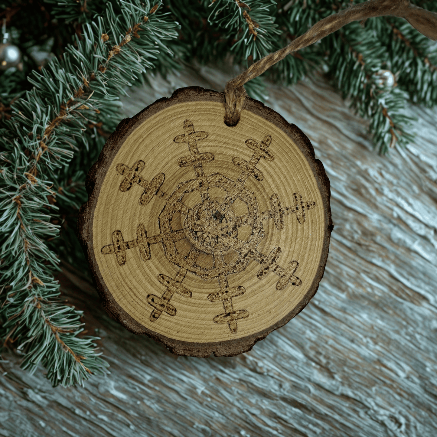 A wooden log slice Christmas ornament on a black background. The ornament is decorated with a detailed snowflake design burned into the wood using pyrography. A jute rope is attached at the top for hanging.