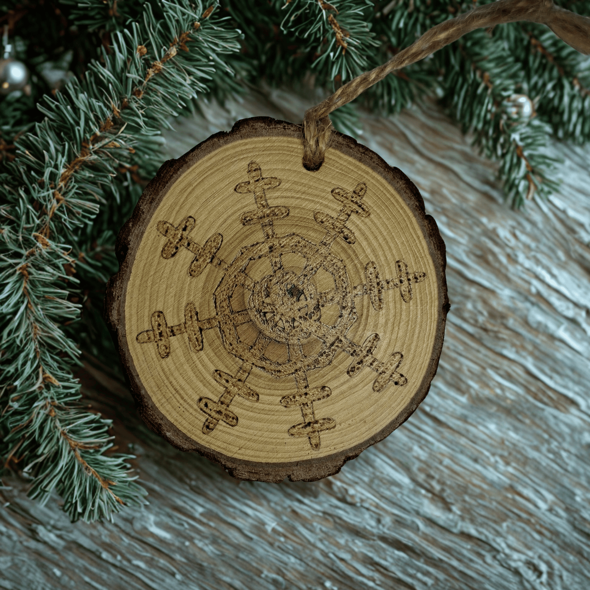 A wooden log slice Christmas ornament on a black background. The ornament is decorated with a detailed snowflake design burned into the wood using pyrography. A jute rope is attached at the top for hanging.