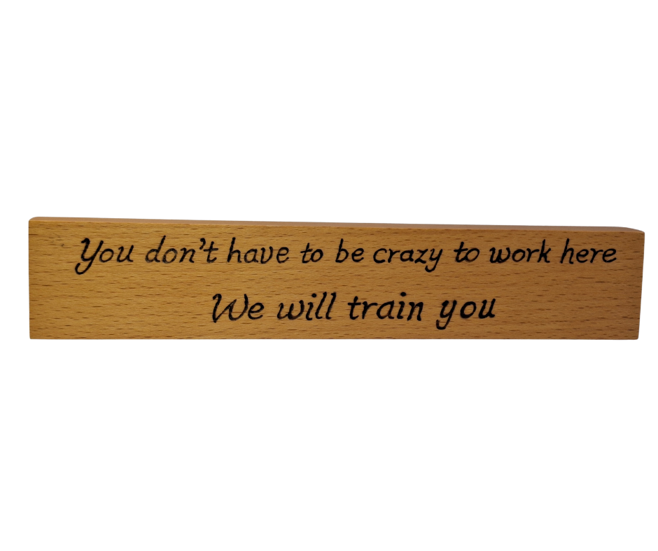 A hand-burnt, freestanding wooden plaque with the words "You don't have to be crazy to work here we will train you" in a casual, handwritten style. The wood has visible grain and a rustic, natural finish.