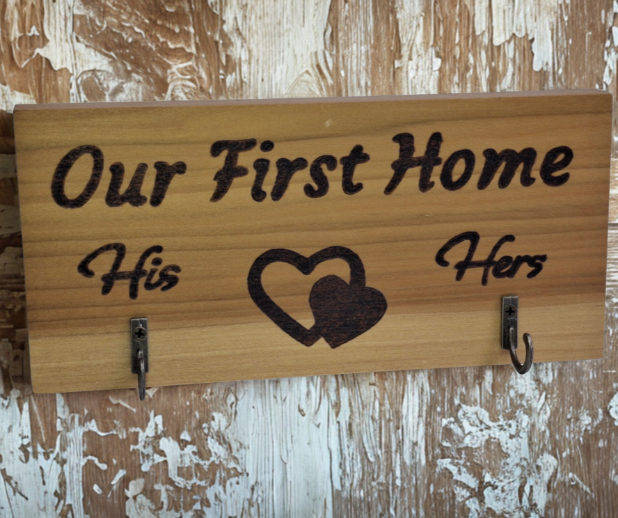 A two-hook hanger crafted from two-tone Tulipwood, featuring hand-burnt lettering that reads "Our First Home" with a double heart motif. The wood has visible grain, showcasing its natural variations. With his and hers hooks.