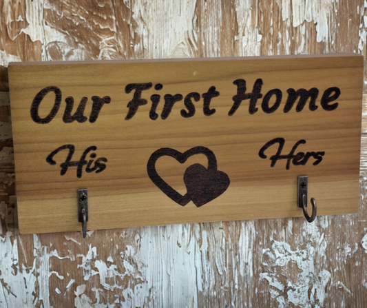 A two-hook hanger crafted from two-tone Tulipwood, featuring hand-burnt lettering that reads "Our First Home" with a double heart motif. The wood has visible grain, showcasing its natural variations. With his and hers hooks.
