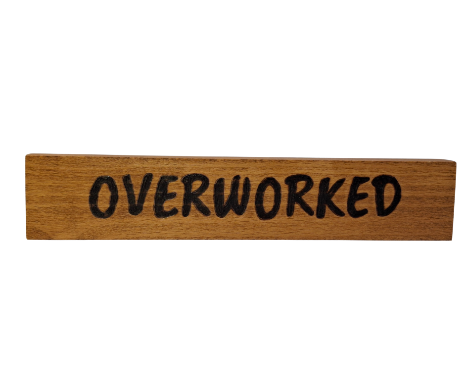 A hand-burnt, freestanding wooden plaque with the word "Overworked" in a casual, handwritten style. The wood has visible grain and a rustic, natural finish.