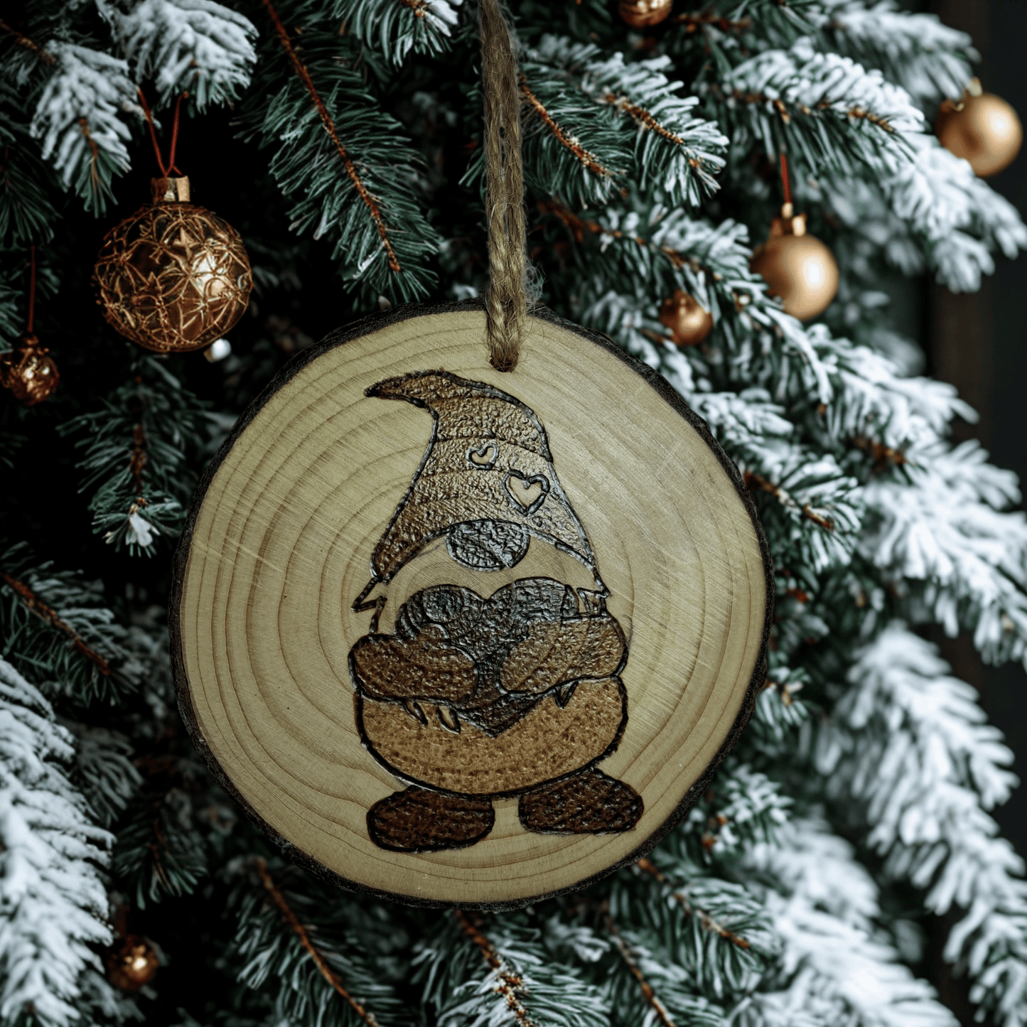 
A small Christmas ornament, a wooden log slice with a jute rope for hanging, is shown on a black background. The ornament is decorated with a pyrography design of a gonk holding a heart, with a pattern of smaller hearts on its hat.