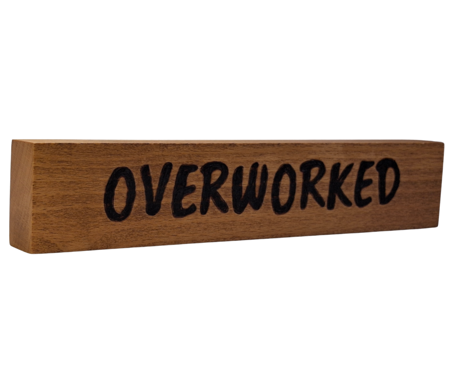 A hand-burnt, freestanding wooden plaque with the word "Overworked" in a casual, handwritten style. The wood has visible grain and a rustic, natural finish.