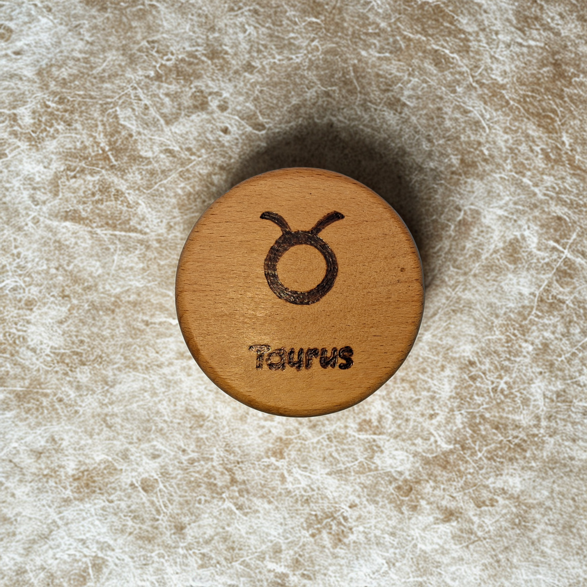 Close-up of a light-colored beechwood circle, 5cm in diameter and 0.4cm thick, featuring the words "Taurus" and an illustration of the star sign symbol,  hand-burned into its surface using pyrography. The wood has a smooth, waxed finish and the text is clearly legible.