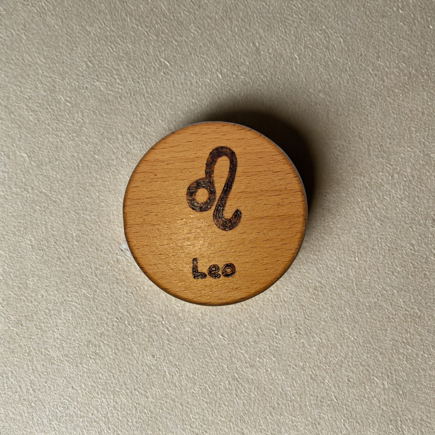 Close-up of a light-colored beechwood circle, 5cm in diameter and 0.4cm thick, featuring the words "Leo" and an illustration of the star sign symbol,  hand-burned into its surface using pyrography. The wood has a smooth, waxed finish and the text is clearly legible.