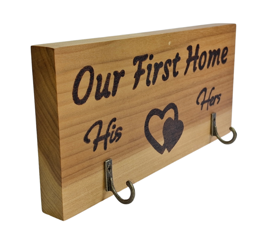 A two-hook hanger crafted from two-tone Tulipwood, featuring hand-burnt lettering that reads "Our First Home" with a double heart motif. The wood has visible grain, showcasing its natural variations. With his and hers hooks.