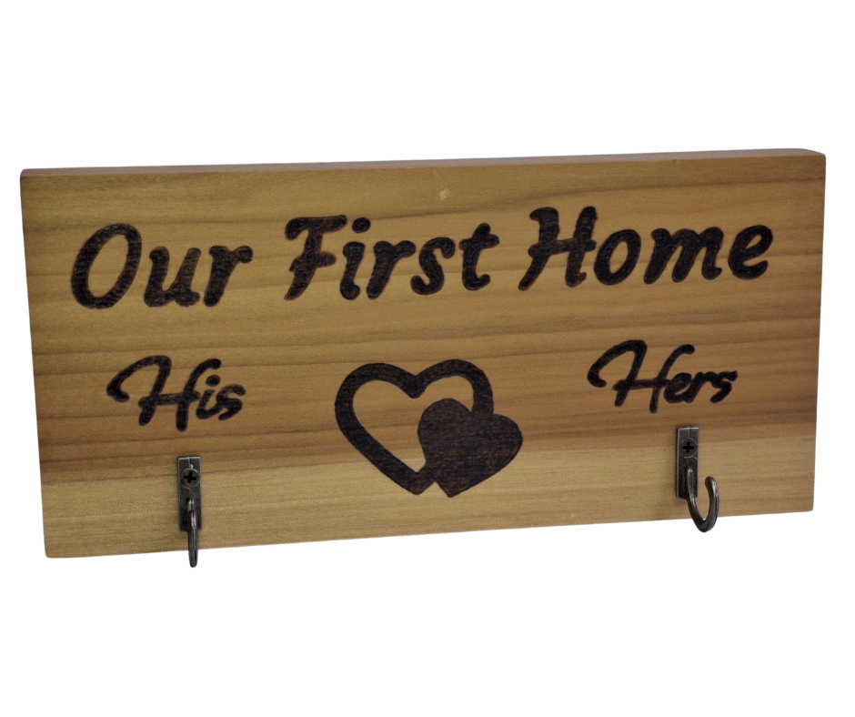 A two-hook hanger crafted from two-tone Tulipwood, featuring hand-burnt lettering that reads "Our First Home" with a double heart motif. The wood has visible grain, showcasing its natural variations. With his and hers hooks.