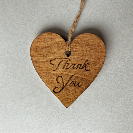 Heart-shaped wooden ornament, hand-drawn with pyrography, featuring the engraved phrase "Thank you" in a decorative script, set against a textured background.