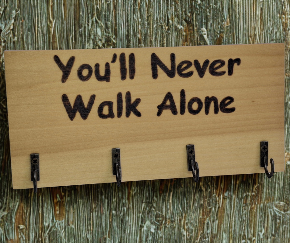 A four-hook hanger crafted from two-tone Tulipwood, featuring hand-burnt lettering that reads "You'll Never Walk Alone." The wood has visible grain, showcasing its natural variations.