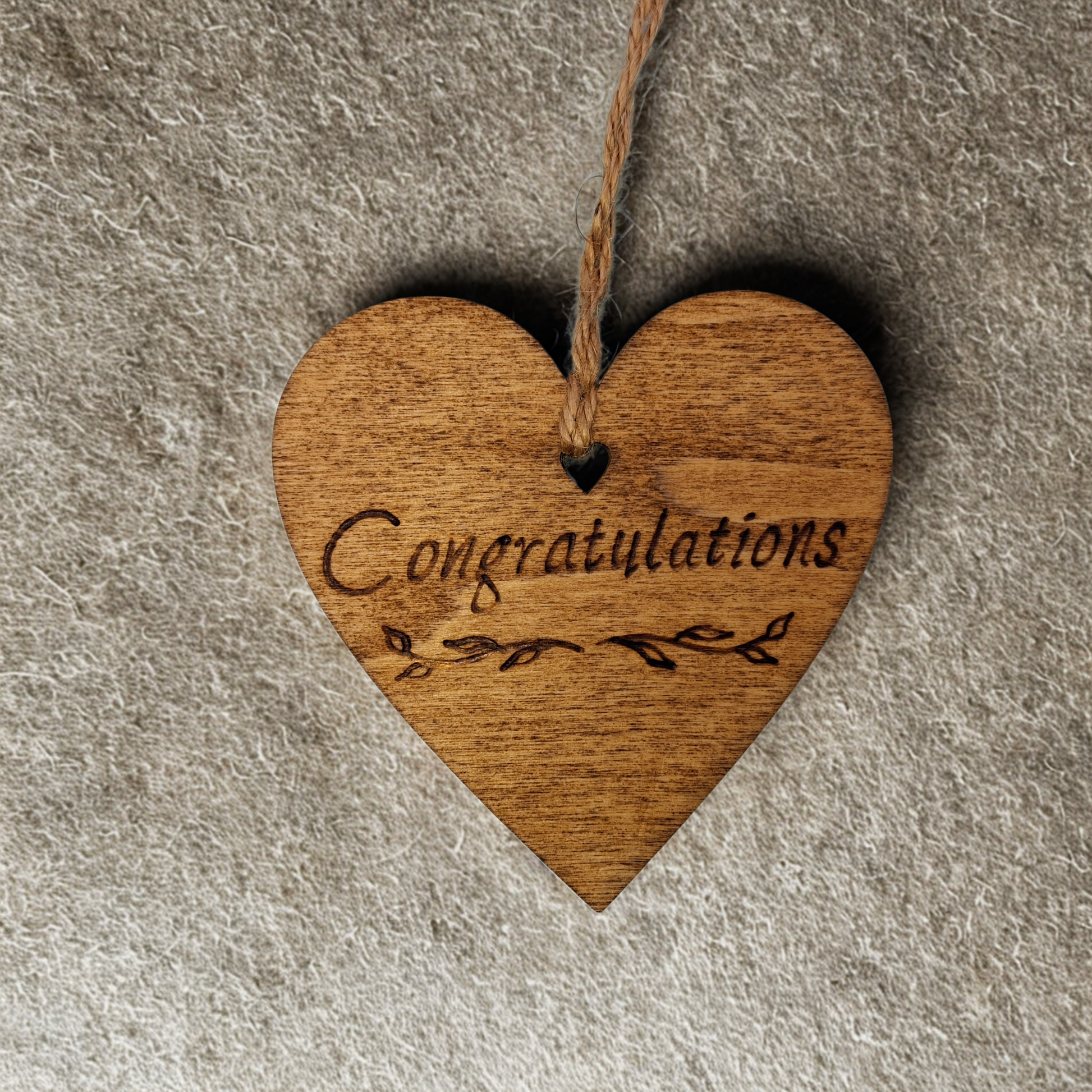 
Heart-shaped wooden ornament, hand-drawn with pyrography, featuring the engraved word "Congratulations" with decorative flourishes, set against a textured background.