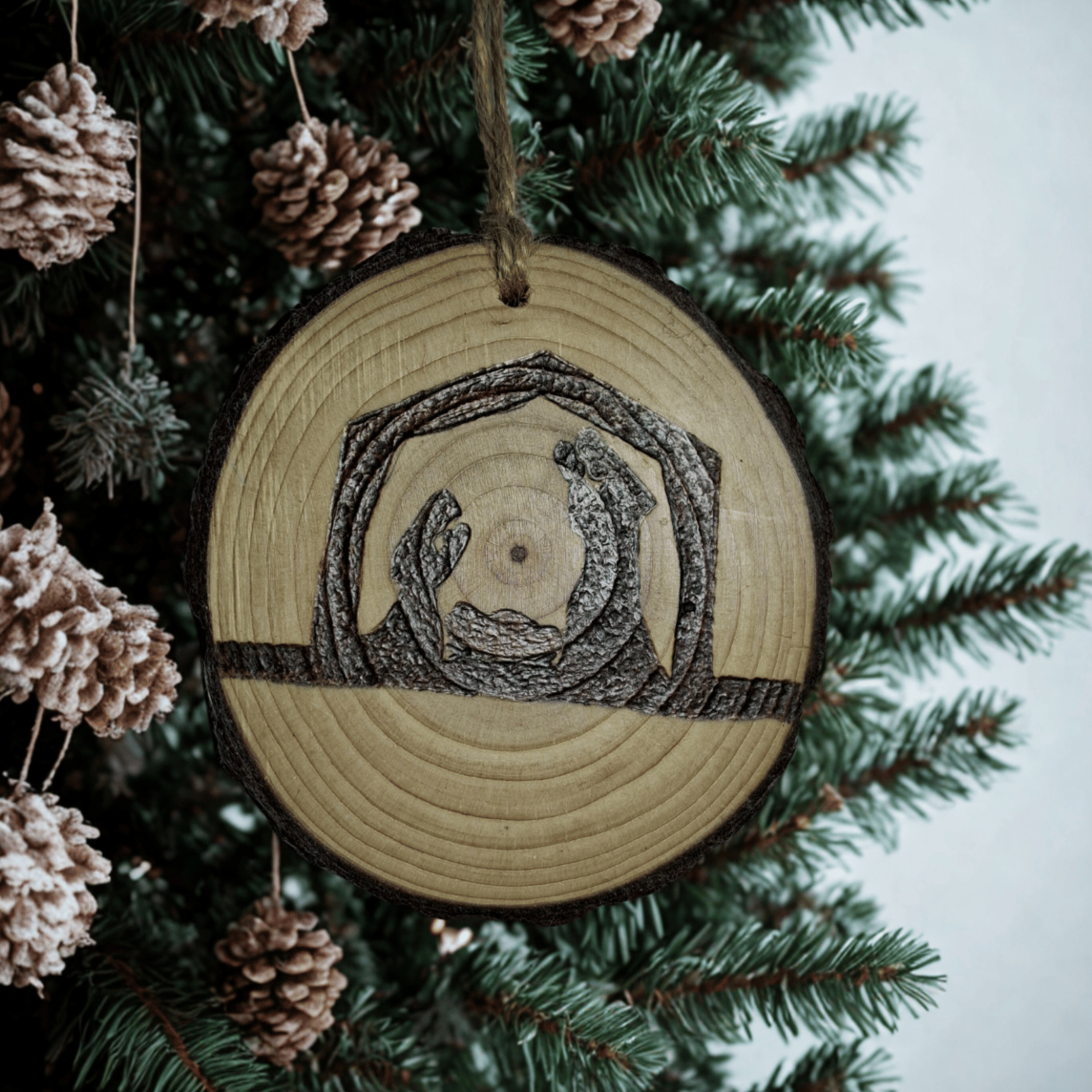 A small Christmas ornament, a wooden log slice with a jute rope for hanging, is shown on a black background. The ornament is decorated with a pyrography design of a nativity scene featuring Mary, Joseph, and baby Jesus in a manger.