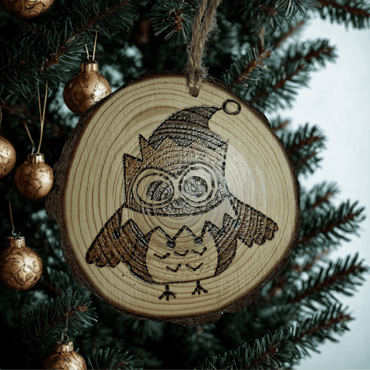 
A close-up of a Christmas ornament on a black background. The ornament is a round wooden log slice with a jute rope for hanging. The front of the ornament is decorated with a pyrography design of a cute owl with large eyes, wearing a pointed hat.