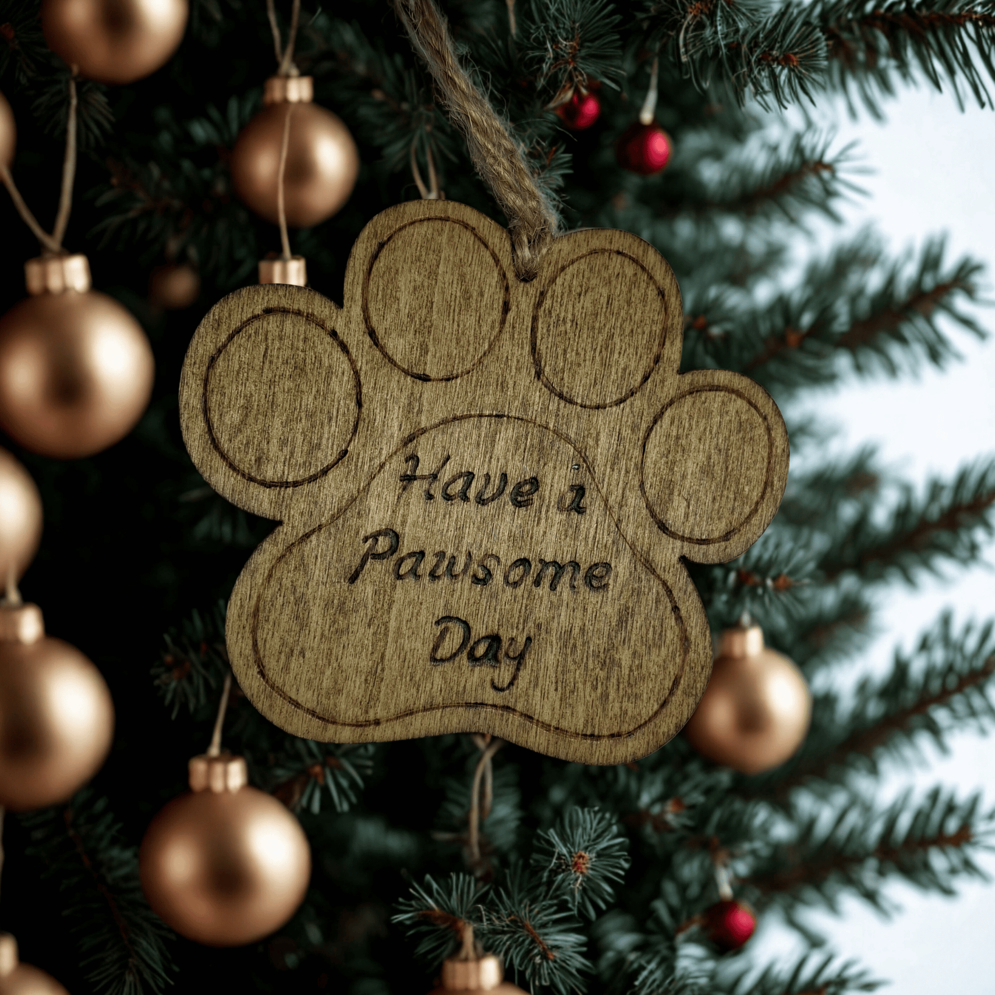 
A light brown, wooden, paw-print-shaped ornament. It has a hole at the top with a piece of natural twine for hanging. The words "Have a Pawsome Day" are hand burnt in a cursive font into the center of the paw. The background is solid black.