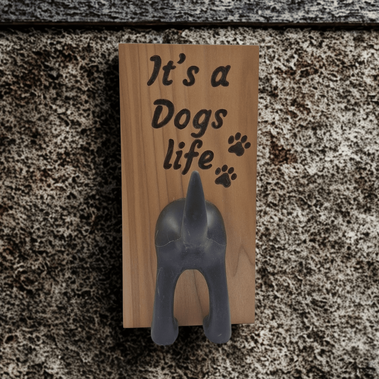 A vertical wooden wall plaque featuring the hand-burnt with pyrography text "It's a Dogs life" in a black, casual script. To the right of the word "life" are three small paw prints. A black, molded figure of a dog's rear end is attached below the text, with the upturned tail forming a hook. 