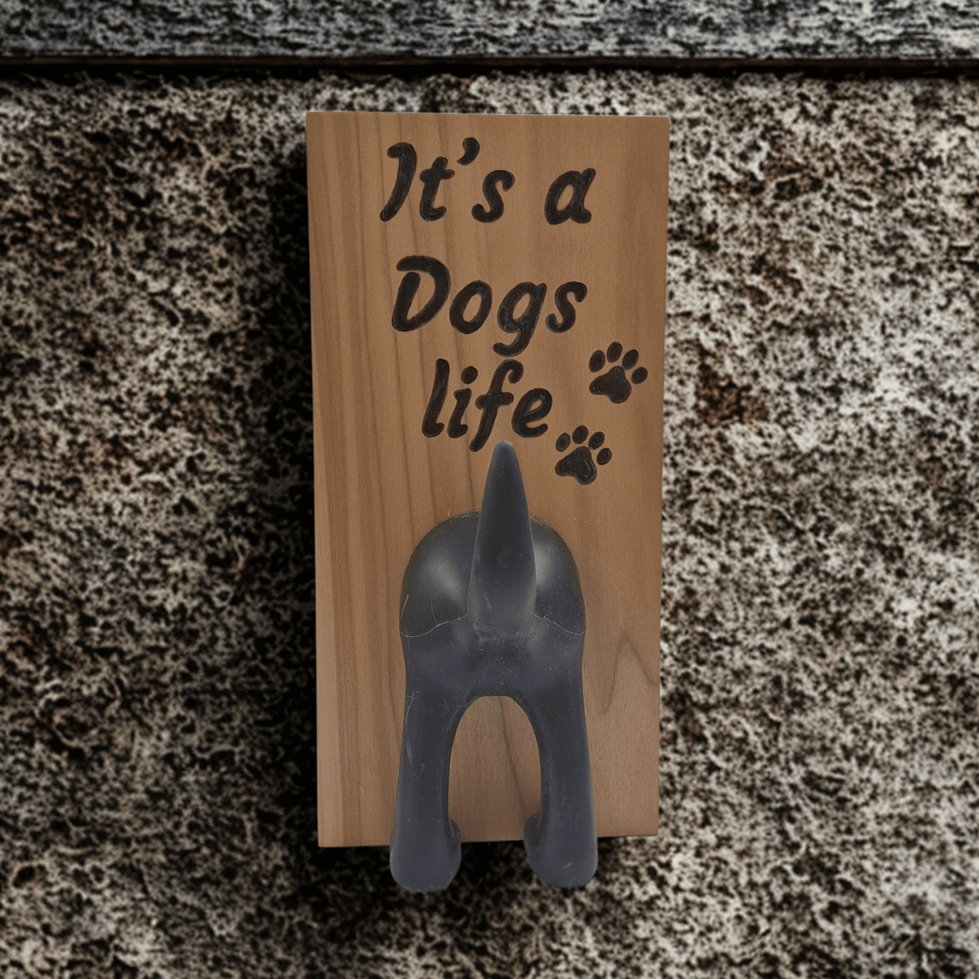 A vertical wooden wall plaque featuring the hand-burnt with pyrography text "It's a Dogs life" in a black, casual script. To the right of the word "life" are three small paw prints. A black, molded figure of a dog's rear end is attached below the text, with the upturned tail forming a hook. 