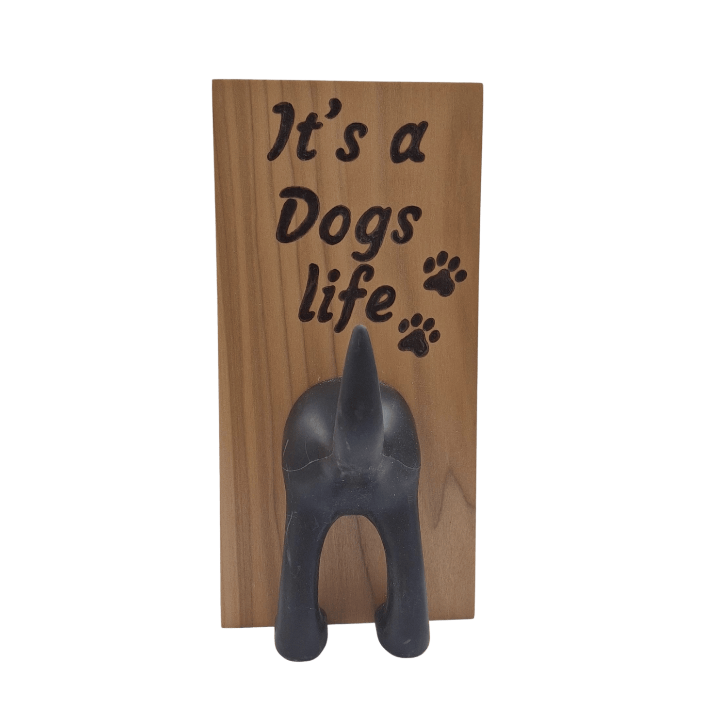 A vertical wooden wall plaque featuring the hand-burnt with pyrography text "It's a Dogs life" in a black, casual script. To the right of the word "life" are three small paw prints. A black, molded figure of a dog's rear end is attached below the text, with the upturned tail forming a hook. 