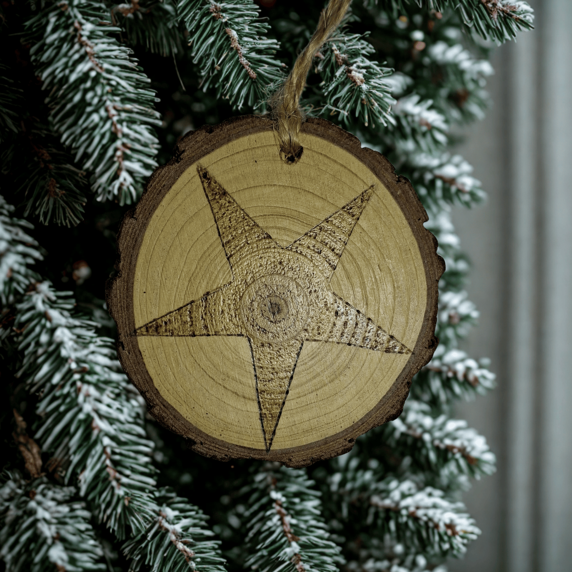 
A close-up of a Christmas ornament on a black background. The ornament is a round wooden log slice with a jute rope for hanging. The front of the log slice is decorated with a pyrography design of a five-pointed star.