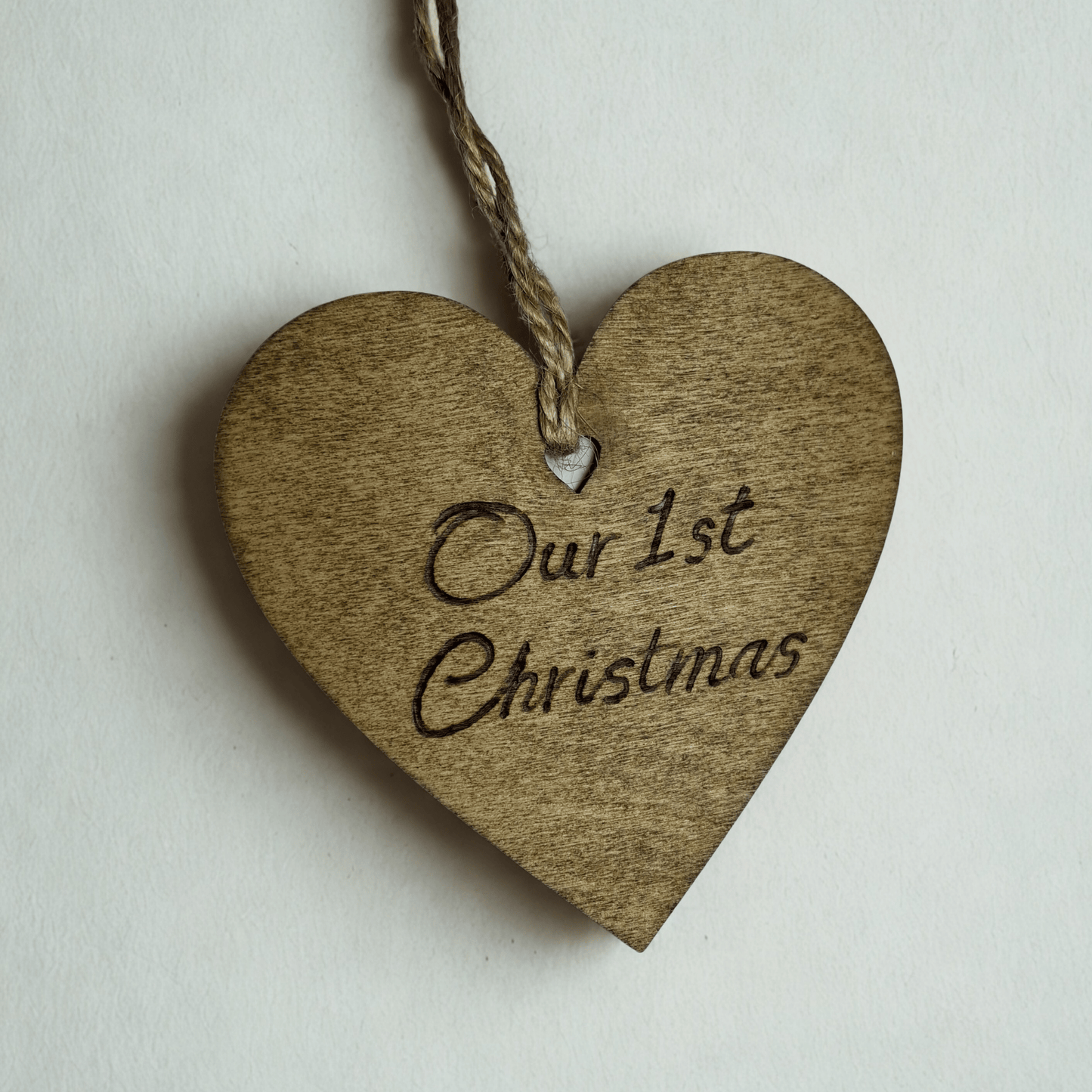 
A close-up of a brown, wooden heart-shaped decoration with a hole at the top. A piece of twine goes through the hole for hanging. The words "Our 1st Christmas" are carved into the front of the heart. The background is solid black.