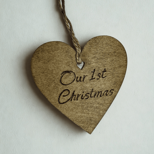 
A close-up of a brown, wooden heart-shaped decoration with a hole at the top. A piece of twine goes through the hole for hanging. The words "Our 1st Christmas" are carved into the front of the heart. The background is solid black.