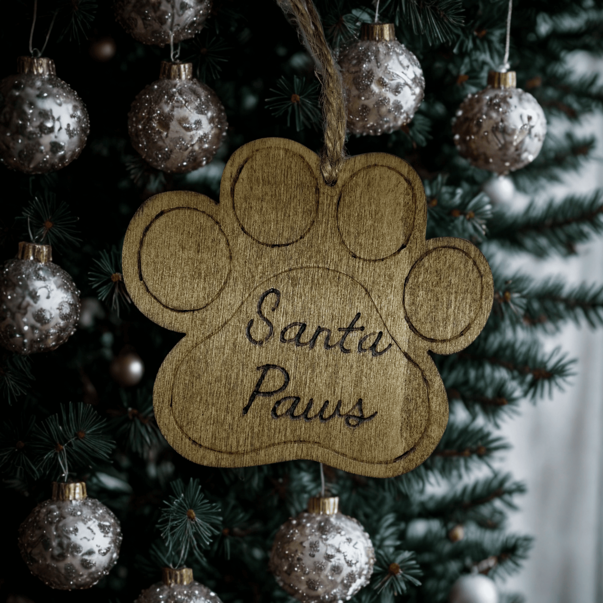 
A light brown wooden decoration in the shape of a dog's paw print. A piece of natural twine is threaded through a hole at the top for hanging. The words "Santa Paws" are carved in a cursive font into the center of the paw. The background is solid black.