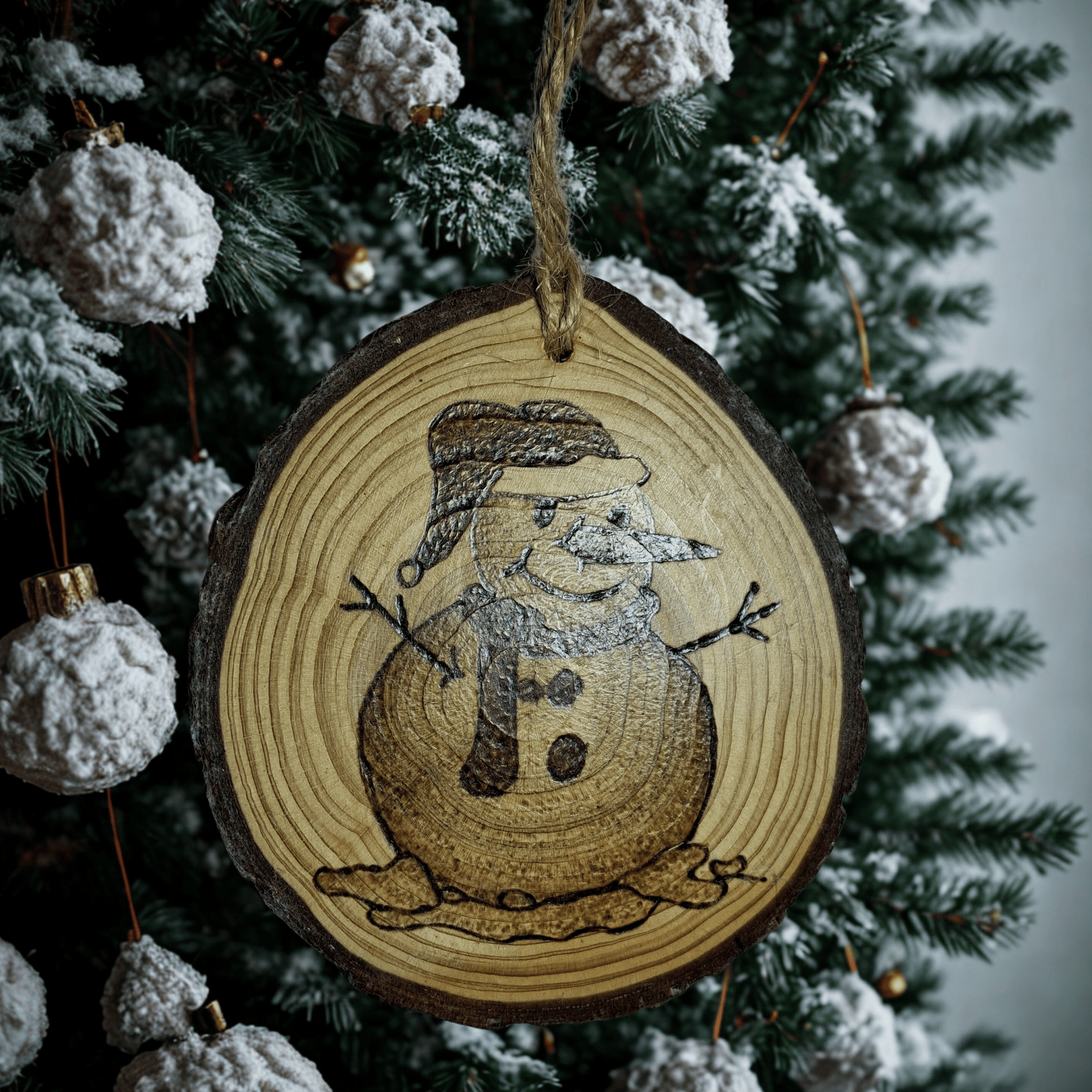 
A Christmas ornament on a black background. The ornament is a round wooden log slice with a jute rope for hanging. It is decorated with a pyrography design of a snowman with a hat and scarf, with snow melting around his base.