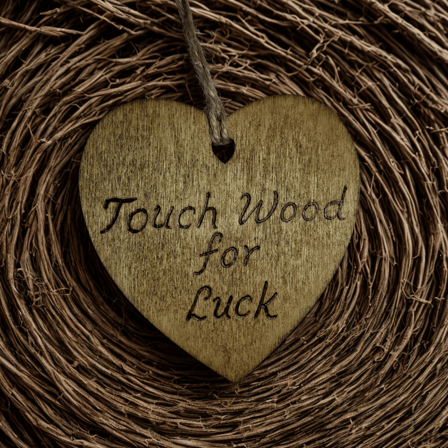 
A brown, wooden, heart-shaped plaque with a hole at the top for a string to hang it. The string is made of twisted twine. The words "Touch Wood for Luck" are burned into the surface in a handwritten font. The heart is lit from the front, making the wood grain visible. The background is solid black.