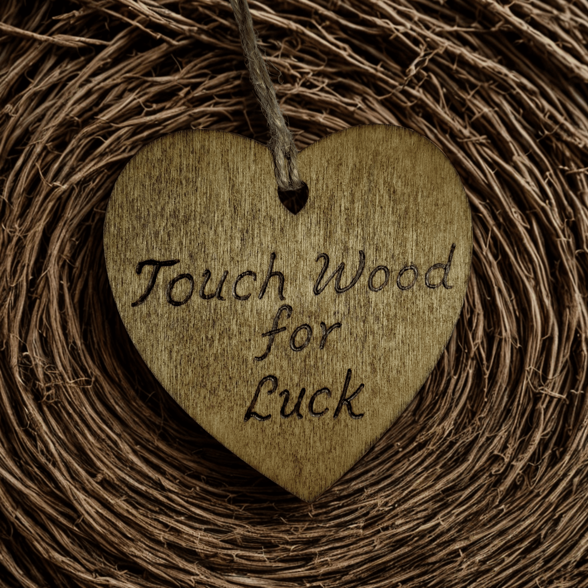 
A brown, wooden, heart-shaped plaque with a hole at the top for a string to hang it. The string is made of twisted twine. The words "Touch Wood for Luck" are burned into the surface in a handwritten font. The heart is lit from the front, making the wood grain visible. The background is solid black.