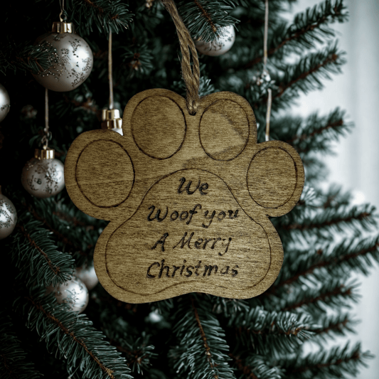 A close-up of a brown, wooden ornament shaped like a dog's paw print. It has a hole at the top with a piece of natural twine for hanging. The words "We Woof you A Merry Christmas" are hand burnt into the center of the paw. The background is solid black.