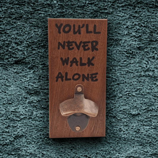 Wall-mounted bottle opener made from dark-grained wood (sapele). The text "YOU'LL NEVER WALK ALONE" is applied using pyrography (hand burnt lettering). A vintage-style metal opener is secured at the bottom of the plaque.